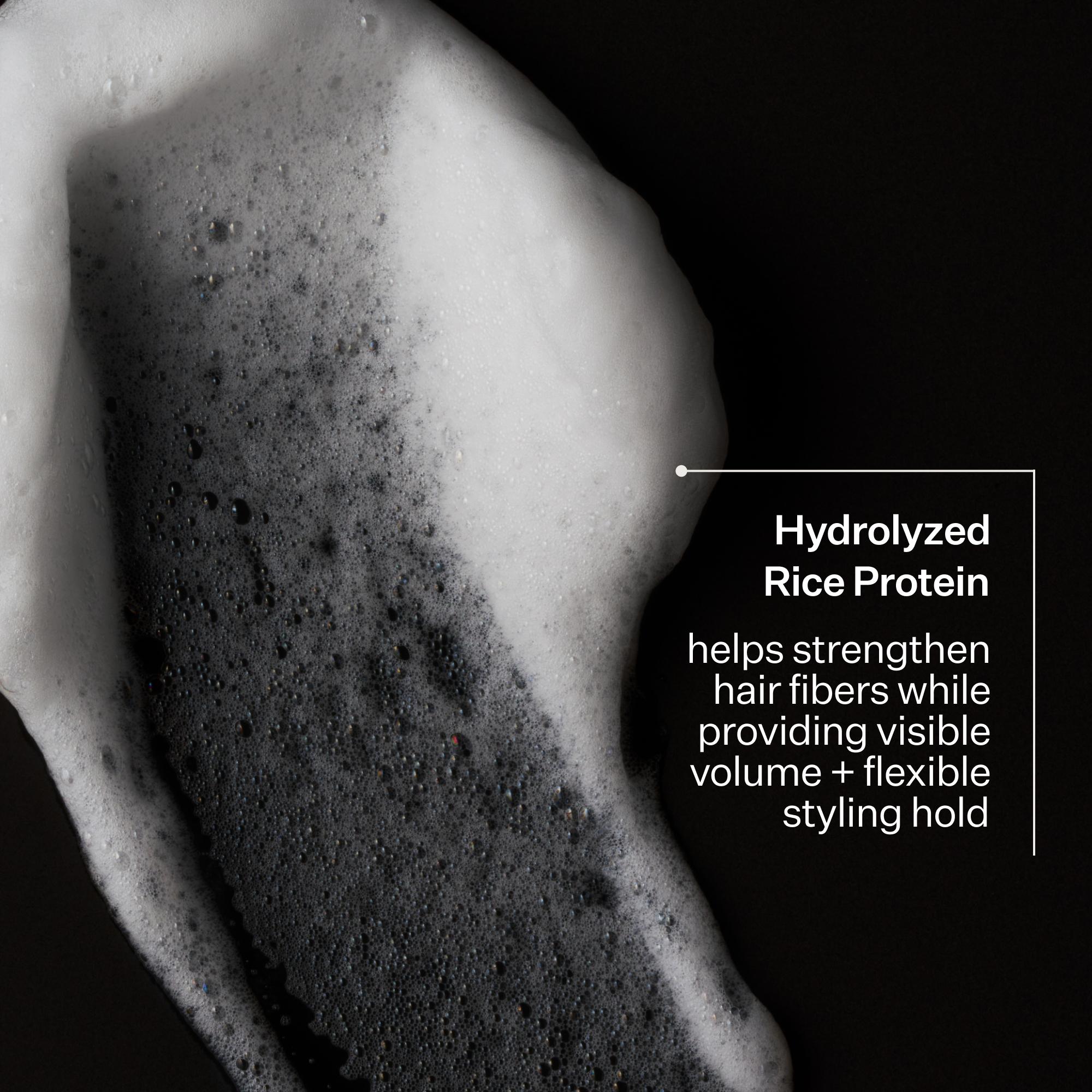 Close-up of white shampoo lather applied to dark hair against a black background, with text reading 'Hydrolyzed Rice Protein' and a descriptive callout.